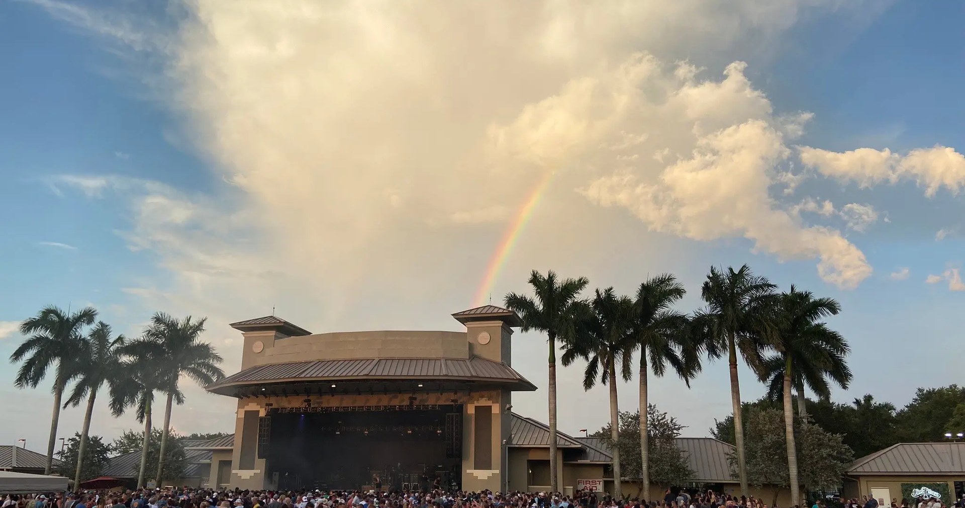 Sunset Cove Amphitheater at golden hour, a rainbow over the stage and royal palms, crowd on the lawn